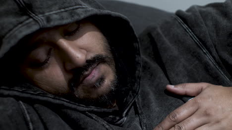 An-Indian-man-wearing-a-hoodie,-eyes-closed-has-he-relaxes-on-a-sofa-with-his-hand-tapping-to-a-rhythmic-beat-on-his-chest