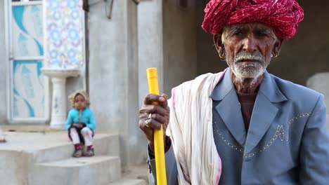 Portrait-of-old-senior-Indian-man-wearing-red-turban-leaning-on-yellow-stick-and-child-sitting-on-stairs-in-background
