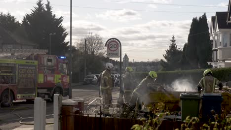 Firefighters-extinguishing-a-smouldering-fire-in-a-skip-bin-outside-a-residential-home,-the-bin-filled-with-building-rubbish-as-the-fire-brigade-attempt-to-neutralise-the-threat,-London,-England