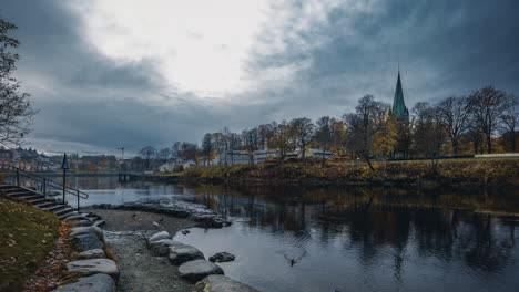 Düsterer-Himmel-über-Dem-Fluss-Nidelva-In-Trondheim,-Norwegen-Mit-Blick-Auf-Die-Kathedrale-Von-Nidaros-Und-Einem-Mann,-Der-Im-Herbst-Enten-Am-Flussufer-Füttert