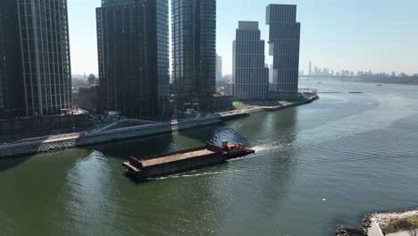 An-aerial-view-of-a-barge-sailing-down-Newtown-Creek-with-new-high-rise-apartment-buildings-in-Brooklyn,-NY-in-the-background-on-a-sunny-day