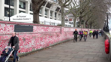 A-man-with-a-pushchair-followed-by-a-group-of-cyclists-pass-the-thousands-of-red-painted-hearts-with-messages-to-lost-loved-ones-during-the-pandemic-at-The-National-Covid-Memorial-Wall