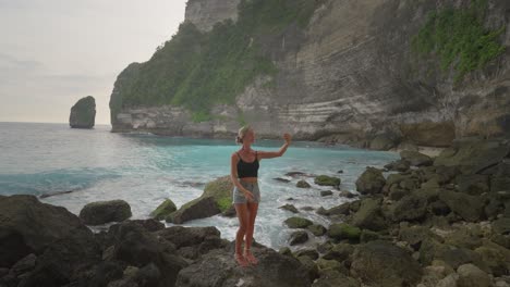 Selfie-taking-woman-on-shore-of-tropical-island-with-grand-cliffs,-Nusa-Penida