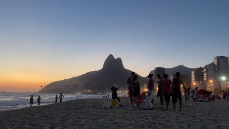 Silhouetted-people-on-the-beach-of-Leblon-with-Two-Brothers-mountain-and-city-lights-of-shantytown-in-the-background-and-leisure-and-sports-activities-in-the-foreground
