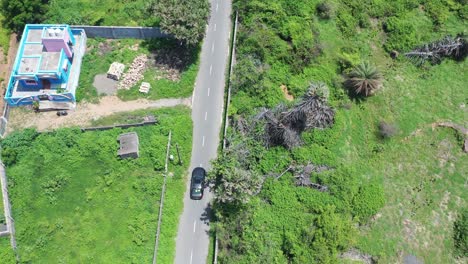 aerial-footage-of-a-black-luxury-car-on-a-concrete-road-through-greenery-and-various-buildings,-cityscape,-empty-roads,-residential-area-with-a-green-background,-coconut-trees,-car-being-followed