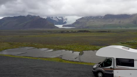 Glaciar-De-Islandia-En-La-Distancia-Con-Dos-Vehículos-Recreativos-En-Primer-Plano-Con-Video-De-Drones-Avanzando