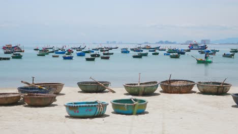 Traditional-Craft-Wooden-Basket-Boats-on-Sandy-Beach-at-Da-Nang-City-Vietnam,-Fishing-Boat-Sailing-Passing-on-Water,-Lot-of-Canoe-and-Small-Boats-Anchored-in-Sea