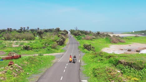 aerial-view-of-man-and-woman-on-two-wheelers-approaching-camera-from-the-other-direction,-bright-blue-clear-sky-in-background,-grass-on-sides-of-road,-carefree-couple,-focus-on-background,-scenic-view
