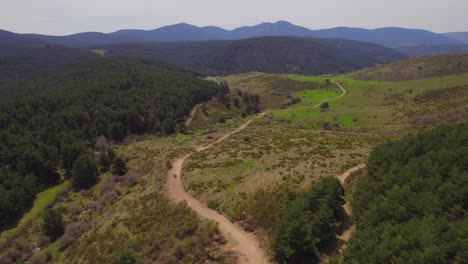 Aerial-drone-shot-of-car-exploring-nature-of-Spain-on-sandy-path-between-green-hills-and-mountain-range-in-backdrop---Beautiful-summer-day-in-Wilderness