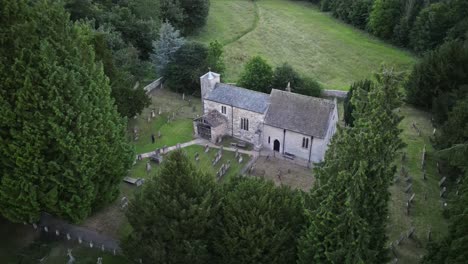 Suave-Arco-De-Drones-Aéreos-Entre-Coníferas-Altas,-Que-Muestra-La-Antigua-Iglesia-En-El-Entorno