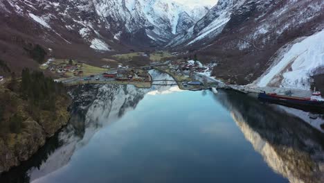 High-altitude-aerial-approaching-amazing-old-Viking-village-Gudvangen-in-Norway---Aerial-looking-down-with-blue-sky-and-mountain-reflections-in-crystal-clear-water-surface-during-winter