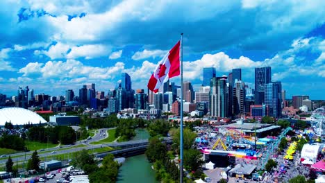 Calgary-Stampede-arial-drone-hold-horizon-city-skyline-sunny-summer-day-largest-Canada-flag-in-the-foreground-MNP-community-sports-Repsol-Centre-divided-by-river-running-lush-green-thousands-of-people