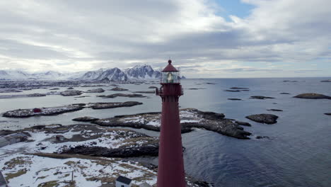 Ariel-horizontal-orbit-of-the-Andenes-lighthouse-with-stunning-views-of-the-rocky-coast-and-snow-covered-mountains,-with-the-light-flashing