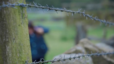 Out-of-focus-walker,-hiker-puting-on-her-rucksack,-shot-through-a-farmland-fence-and-stone-wall