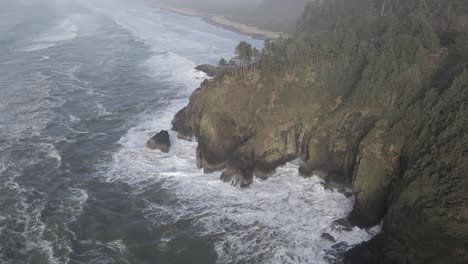 Ocean-waves-crash-against-the-rugged-forested-coastline,-misty,-aerial