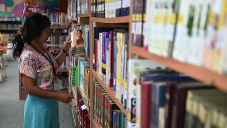 Asian-woman-approaches-the-bookshelves-to-grab-a-book-as-she-giggles-finding-her-favorite-book