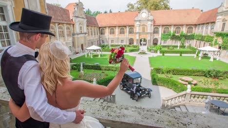 Ein-Glückliches-Brautpaar-Steht-Auf-Dem-Balkon-Eines-Schlosses-Und-Blickt-Auf-Ein-Stilvolles-Auto