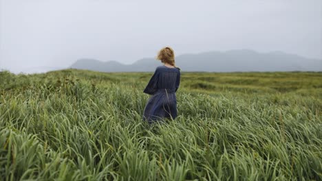 Blond-caucasian-woman-standing-backwards-alone-and-looking-into-the-horizon-in-a-green-grass-field-prairie-during-a-windy-day