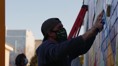 Man-in-a-face-mask-helps-paint-a-large-mural-on-the-wall-at-Artsapalooza