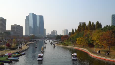 Lake-in-Songdo-Central-Park-in-Incheon-on-sunset,-people-enjoy-traveling-with-paddle-boats-and-walking-along-walk-trails-with-Autumn-city-Skyline