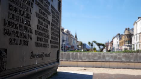 Stadthäuser-Am-Meer-Von-Llandudno-Hinter-Einer-Gedenktafel-Mit-Namen,-Nahaufnahme-Des-Kriegsdenkmals-An-Der-Sonnigen-Promenade,-Dolly-Rechts