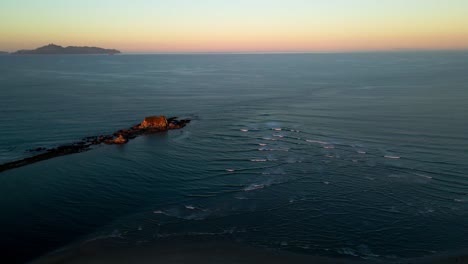 Aerial-flyover-small-waves-of-Pacific-Ocean-with-outstanding-rocks-and-beautiful-colorful-sunset-at-horizon-in-New-Zealand