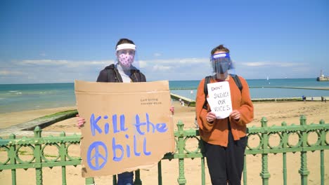 Two-participants-of-a-protest-by-the-seaside-wearing-face-covering-masks-and-holding-cards-with-sea-and-lighouse-in-the-backround