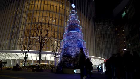 Geschmückter-Weihnachtsbaum-Mit-Bunten-Lichtern-Vor-Dem-Lotte-World-Tower,-An-Dem-Am-Vorabend-Des-Feiertags-In-Der-Nacht-Last-Minute-Käufer-Vorbeilaufen