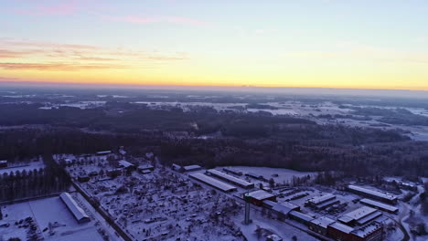 Aerial-backward-moving-shot-over-urban-landscape-with-snow-covered-roofs-and-smoke-coming-out-of-an-industrial-chimney-with-godowns-all-around-at-sunrise