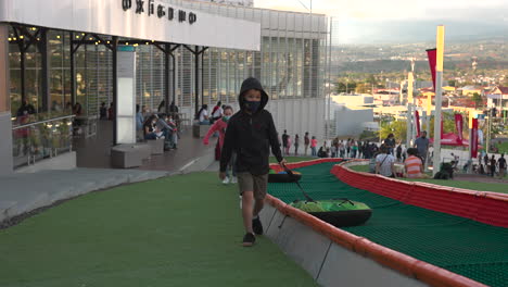 children-running-with-tubby-tire-to-slide-on-green-grass-downhill,-outdoors