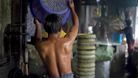 Back-view-of-shirtless-man-pouring-soybeans-into-press-machine-to-make-it-liquid-in-traditional-tofu-factory,-Magelang-in-Indonesia