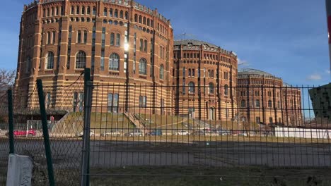 Vienna-gasometer-from-the-side-behind-a-fence-and-a-field-in-the-sunshine,-Austria,-Europe,-moving-shot
