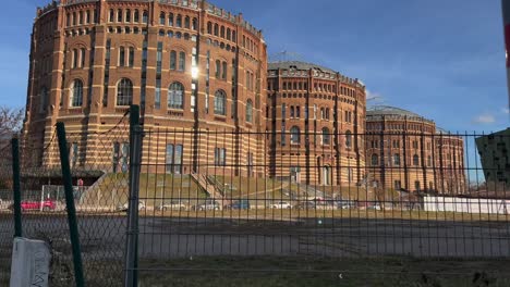 Gasómetro-De-Viena-Desde-El-Lado-Detrás-De-Una-Valla-Y-Un-Campo-Bajo-El-Sol,-Austria,-Europa,-Toma-En-Movimiento