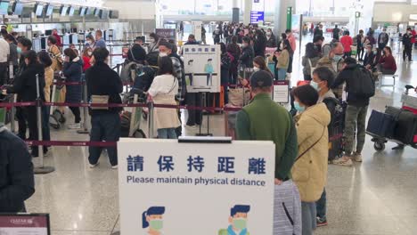 Passagiere-Warten-In-Der-Schlange-Vor-Einem-Check-in-Schalter-Einer-Fluggesellschaft,-Während-Ein-Schild-Die-Menschen-Daran-Erinnert,-Soziale-Distanz-Am-Internationalen-Flughafen-Chek-Lap-Kok-In-Hongkong,-China,-Einzuhalten