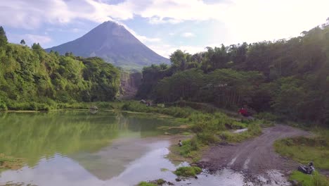 Aerial-view-of-natural-lake-and-driving-old-truck-on-pebbly-road-in-bego-pendem,Indonesia---Beautiful-forest-landscape-and-MERAPI-VOLCANO-in-background