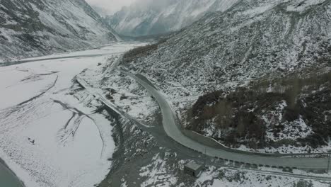 Aerial-Flying-Over-Empty-Road-In-Snow-Covered-Hunza-Valley-Landscape-Beside-Frozen-River