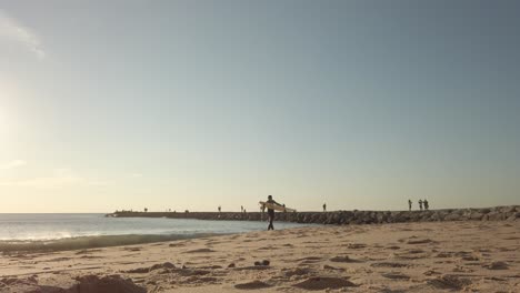 Single-surfer-carrying-board-walking-across-golden-sandy-Portugal-beach-at-sunrise