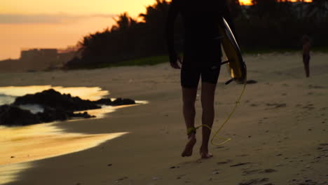 Slow-mo,-surfer-guy-in-wetsuit-holding-surfboard-walking-on-beach-during-golden-hour