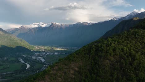 A-dynamic-aerial-shot-moving-over-a-mountain-going-towards-the-towns-of-Villadossola,-Domodossola,-and-the-Simplon-pass-surrounded-by-mountains-and-close-rivers