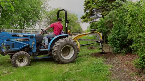 Drilling-holes-for-fence-foundation-with-small-blue-tractor,-farmstead-backyard