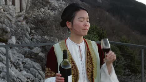 Alegre-Mujer-Asiática-Con-Traje-Tradicional-Montenegrino-Bebiendo-Vino-Tinto-De-Una-Copa-De-Vino-Mientras-Disfruta-De-La-Vista-De-La-Bahía-De-Kotor-En-Sus-Vacaciones