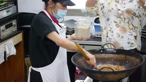 Female-Asian-chef-wearing-face-mask-adds-powder-flavor-into-pot-of-fried-meat-mixture-then-stirs-ingredients-with-spatula-inside-large-wok-with-steam-rising