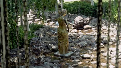 Estatua-Dorada-De-Una-Dama-En-Un-Arroyo-De-Rocas-Y-Una-Fuente-De-Agua-En-Cascada