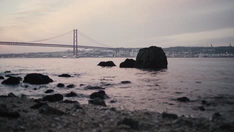 Tagus-river-shore-rocks-waving-with-25th-April-Bridge-at-sunset