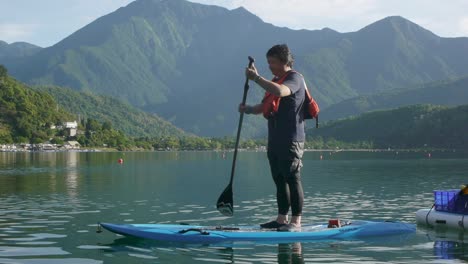 Hombre-Asiático-En-Stand-Up-Paddle-Board-Remando-A-Través-Del-Lago-Rodeado-De-Montañas
