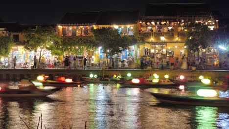 Timelapse-Night-View-of-Boats-Canoe-Traffic-on-River,-illuminated-Floating-Sailing-and-Passing-Fast-at-Lantern-Festival-in-Hoi-An-Vietnam,-Tourists-Nightlife-Bars-and-Cafes-in-Background-on-Quay