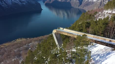Breathtaking-Segastein-viewpoint-high-altitude-aerial-overview-with-person-standing-on-edge---Rotating-around-viewpoint-to-reveal-fjord-and-Aurland-village-in-background-far-down-below