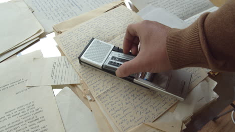 A-man's-hand-inserts-a-microcassette-into-a-retro-handheld-tape-recorder-placed-on-a-stack-of-documents-and-letters