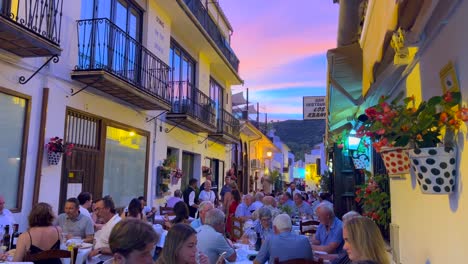 People-sitting-and-having-dinner-outside-in-a-restaurant-with-a-beautiful-sunset-pink-sky-in-Benahavis-Spain,-enjoying-a-lovely-street-in-the-evening-during-summer,-4K-shot