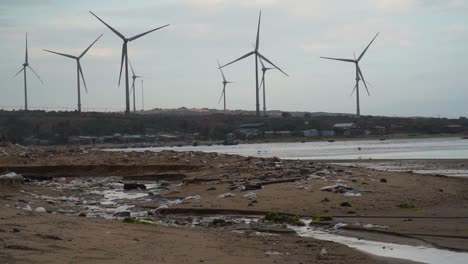 Rubbish-Coastline-At-The-Beach-In-Son-Hai,-Vietnam-With-Wind-Turbines-Spinning-In-Background